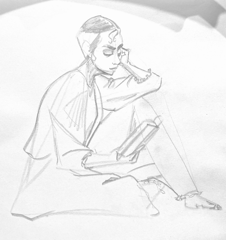 Figure clothed with long sleeves, trousers, holding a book, looking down and reading. Arm resting on their knee.
