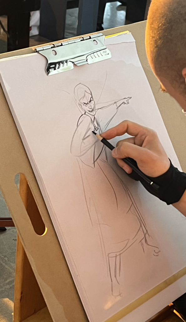 Our guest artist, David Cardenas, drawing with his right hand, of a figure with a snarled face, pointing to the right.