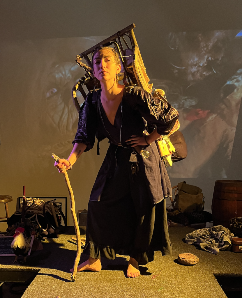 Aslan Scardina posed with hand on hip, hand holding a short wooden staff, carrying a huge backpack of junk sticking out of it. Aslan looks to the right. Aslan stands on the stage, lit with golden yellow light, and a projection of Labyrinth's "Junkyard Lady" in the back.